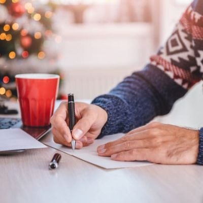A person in a festive sweater writing holiday notes in a foreign language at a desk with a red coffee cup, a laptop and a glowing Christmas tree in the background - it captures the cozy spirit of Christmas.