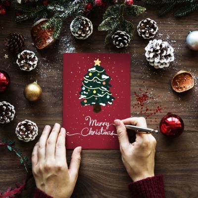 Hände, die „Frohe Weihnachten“ auf eine festliche Karte schreiben, umgeben von Tannenzapfen, Christbaumkugeln und Grünzeug - das fängt den Geist der Feiertage und die mehrsprachige Unterstützung von Skrivanek Baltic ein.
