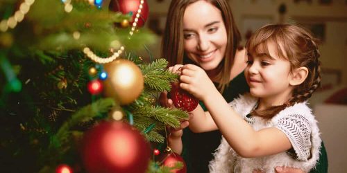 20 funny and entertaining games to celebrate Christmas | Skrivanek Baltic Mother and daughter decorating a Christmas tree with red and gold ornaments, symbolizing festive traditions and togetherness supported by Skrivanek Baltic’s multilingual services.