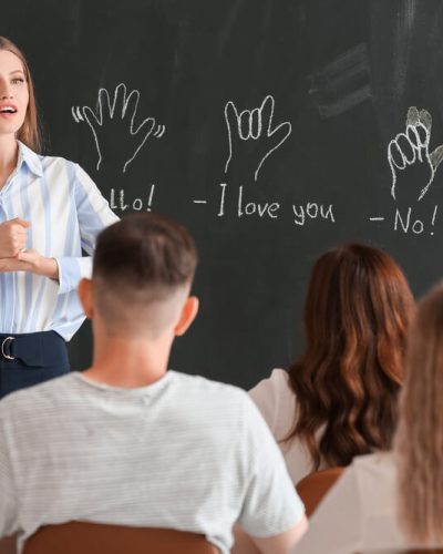 Pasniedzēja māca zīmju valodu studentiem klasē, fonā uz tāfeles parādīti roku žesti ar tulkojumiem – video satura pielāgošana Piekļūstamības direktīvai.