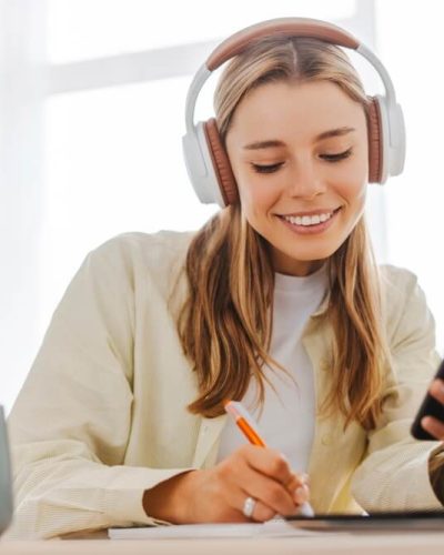 Jauna sieviete ar austiņām smaida, skatoties telefonā un pierakstot idejas pie klēpjdatora – simbolizē radošu tekstu veidošanu un rēgu rakstniecību.