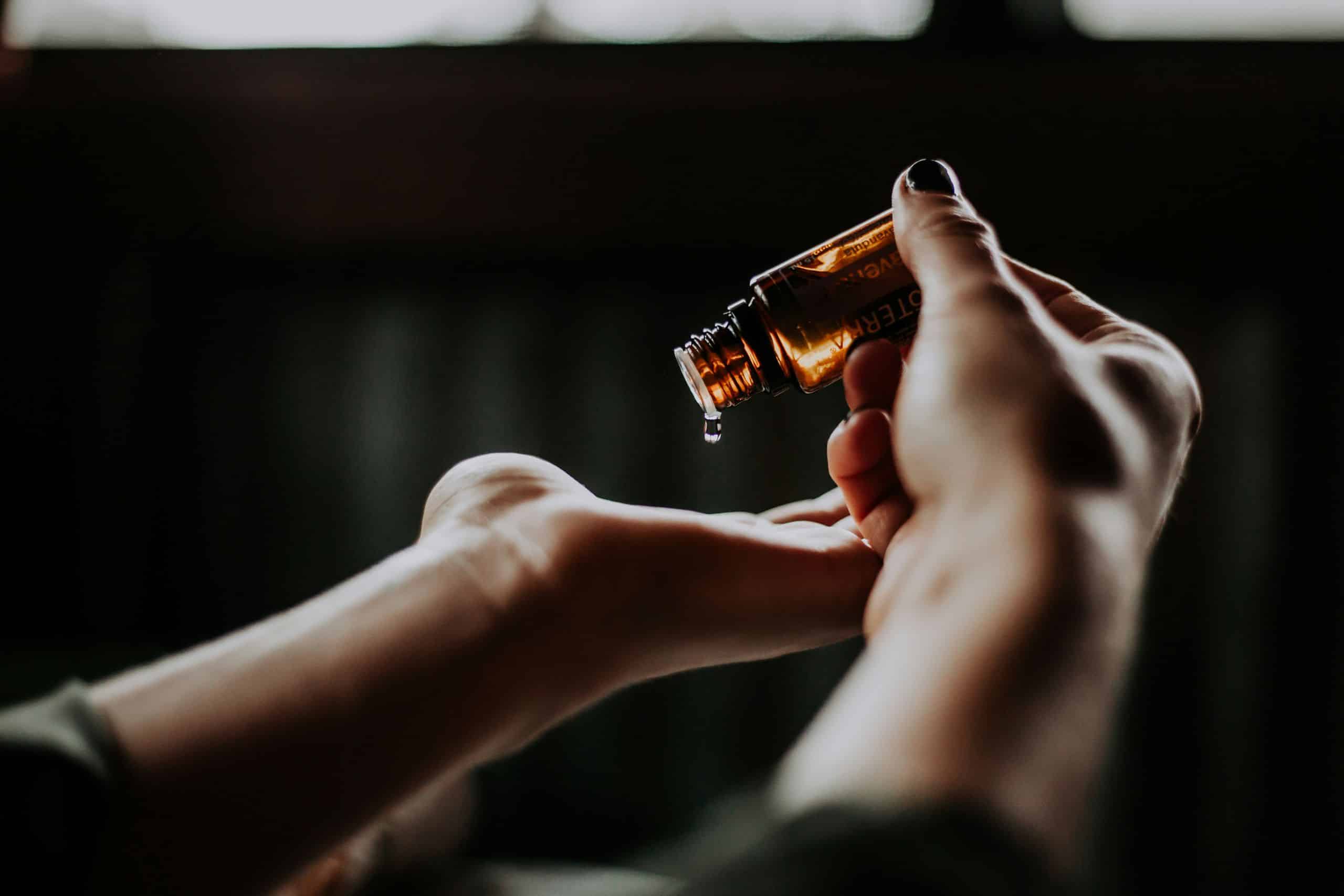 Kāpēc Latvijas Premium SPA Pieredze Pārspēj Ārzemju Kūrortus Cilvēks tur nelielu brūna stikla pudelīti ar ēterisko eļļu un uzmanīgi pilina eļļu plaukstā, fonā redzams vāji izgaismots interjers, radot mierīgu un relaksējošu noskaņu, paspilgtinot kāpēc Latvijas Premium SPA pieredze pārspēj ārzemju kūrortus.