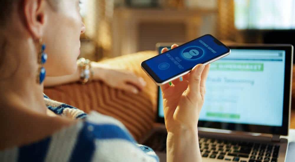 Sieviete dīvānā tur telefonu ar ieslēgtu balss vadības aplikāciju, fonā redzams portatīvais dators.