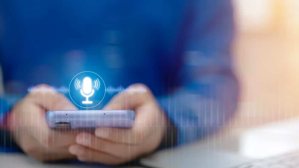 Rokas tur viedtālruni ar aktivizētu balss ierakstīšanas funkciju; redzama mikrofona ikona un digitālie skaņas viļņi.