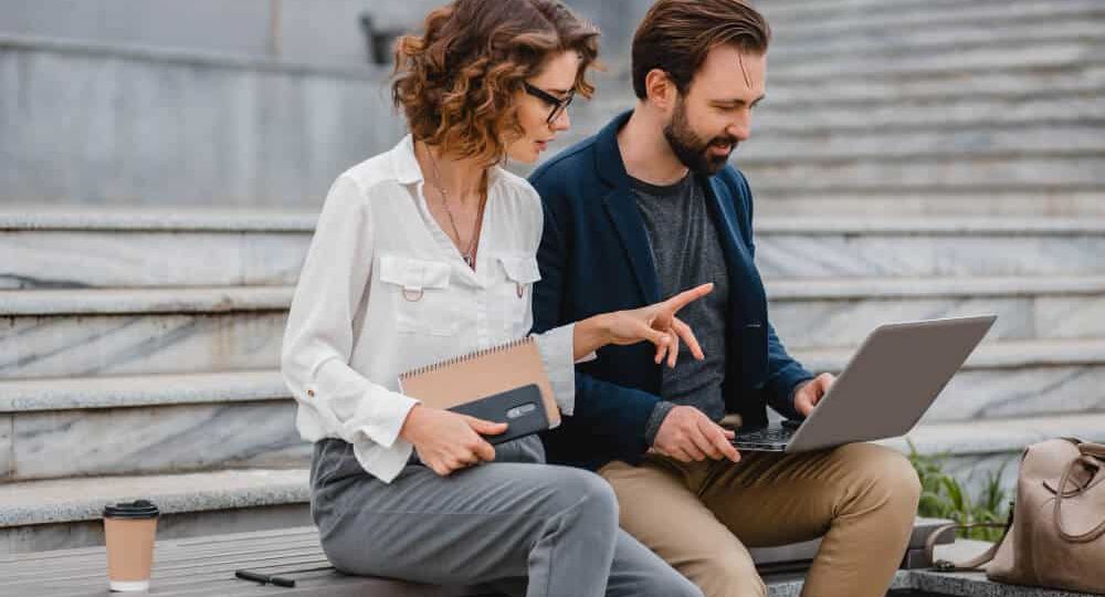 Divi profesionāļi strādā ar klēpjdatoru pilsētvidē, apspriežot digitālus risinājumus un interaktīvās platformas