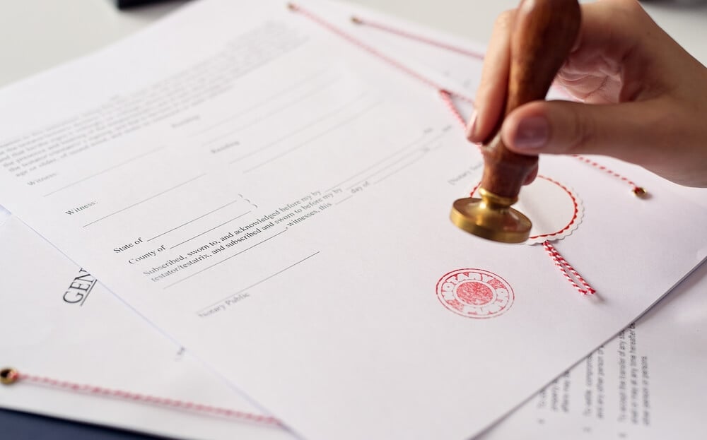 Notariāls tulkojums – kad nepieciešams un kā tas ietekmē cenu Roku tuvplāns ar zīmogu, kas apliecina oficiālu dokumentu – notariāls tulkojums ar sarkanu zīmogu un diegu piesiešanu.
