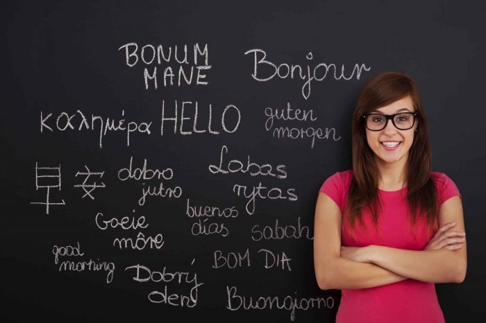 Valodu daudzveidība un tulkošanas māksla – kā saglabāt tēla balsi tulkojumā Sieviete stāv pie tāfeles ar dažādu valodu sveicieniem, piemēram, “Hello”, “Bonjour” un “Guten Morgen”, simbolizējot valodu apguvi un kultūru daudzveidību.