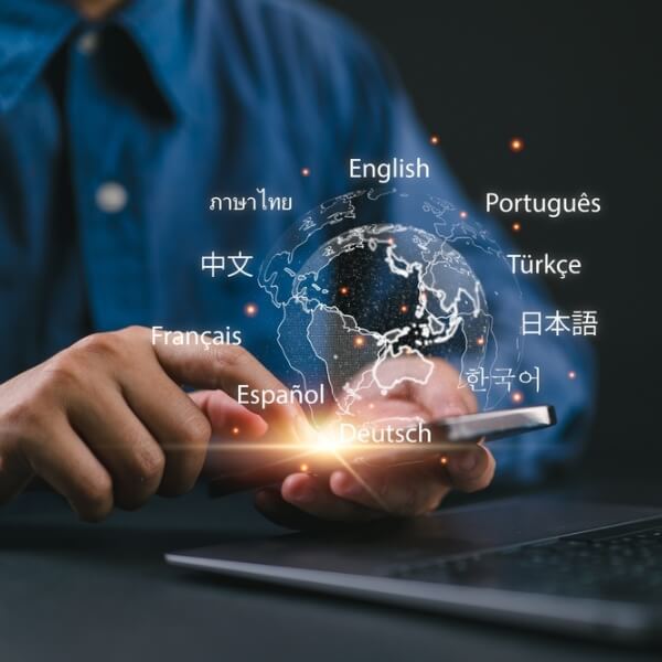 Persona tur viedtālruni virs klēpjdatora, virs pasaules kartes attēloti dažādu valodu nosaukumi, kas simbolizē globālu daudzvalodu digitālo komunikāciju