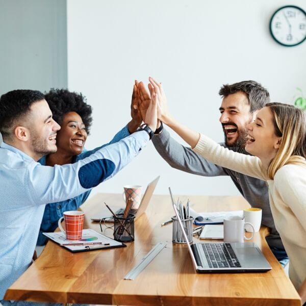 Daudzveidīga komanda birojā sēž pie galda ar klēpjdatoriem un dod piecinieku, atzīmējot veiksmīgu sadarbību un kopīga mērķa sasniegšanu