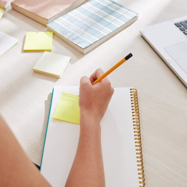 Cilvēks raksta piezīmes kladē pie darba galda, blakus redzami līmlapiņas, grāmatas un klēpjdators, koncentrējoties uz satura formulēšanu