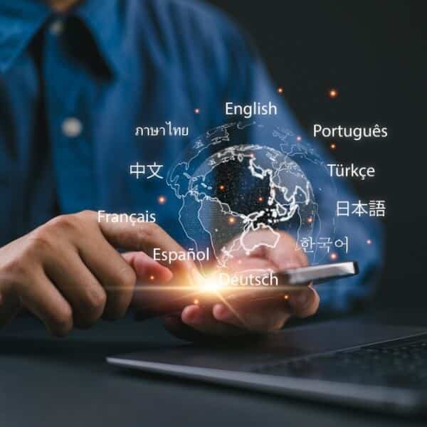 Persona tur viedtālruni virs klēpjdatora, virs pasaules kartes attēloti dažādu valodu nosaukumi, kas simbolizē globālu daudzvalodu digitālo komunikāciju