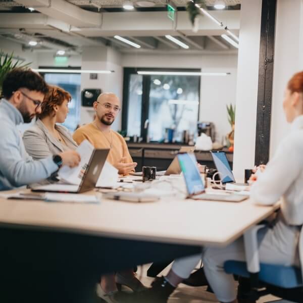 Starptautiska komanda sēž pie kopīga galda modernā birojā, strādā ar klēpjdatoriem un dokumentiem, apspriežot satura un komunikācijas stratēģiju dažādām valodām