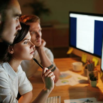 Trīs speciālisti strādā pie datora ekrāna, analizējot un rediģējot digitālu saturu daudzvalodu vidē – komanda koncentrējas uz starptautisko izaugsmi un lokalizāciju, pārvaldot tulkošanas procesus, kvalitātes kontroli un cilvēka kontrolētu lokalizāciju profesionālā darba vidē