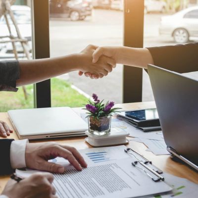 Biznesa tikšanās ar rokasspiedienu pie darba galda, simbolizējot uzticamu sadarbību mašīntulkošanas, pēcrediģēšanas un profesionālu tulkojumu kvalitātes nodrošināšanā